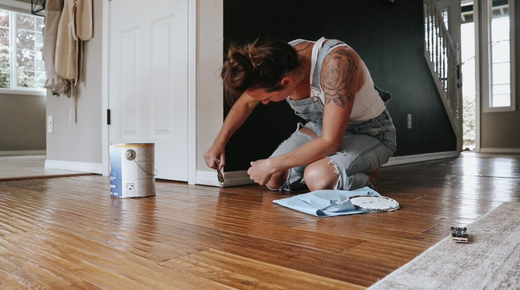 christian homemaker wearing overalls an a white tank top is painting the basesboards in her house